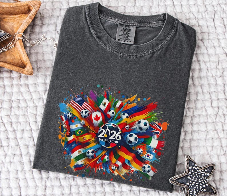 May include: Dark gray t-shirt featuring a colorful graphic design for the 2026 World Cup. The design includes soccer balls and flags from various countries, with the text "2026 WORLD CUP" in the center.