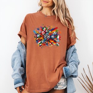 May include: A rust-colored t-shirt featuring a colorful graphic design for the 2026 World Cup. The design includes soccer balls, flags from various countries, and the text "2026 World Cup" in the center.