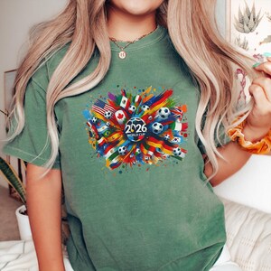 May include: Green t-shirt featuring a colorful graphic design for the 2026 World Cup. The design includes soccer balls, flags from various countries, and the text "2026 WORLD CUP" in white.