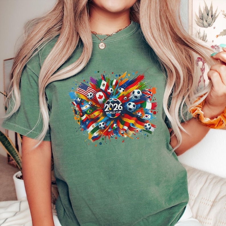 May include: Green t-shirt featuring a colorful graphic design for the 2026 World Cup. The design includes soccer balls, flags from various countries, and the text "2026 WORLD CUP" in white.