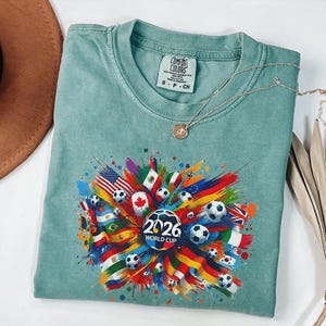 May include: A light teal Comfort Colors t-shirt featuring a colorful graphic design for the 2026 World Cup. The design includes soccer balls, flags from various countries, and the text "2026 WORLD CUP". A silver necklace with a pendant is also visible.