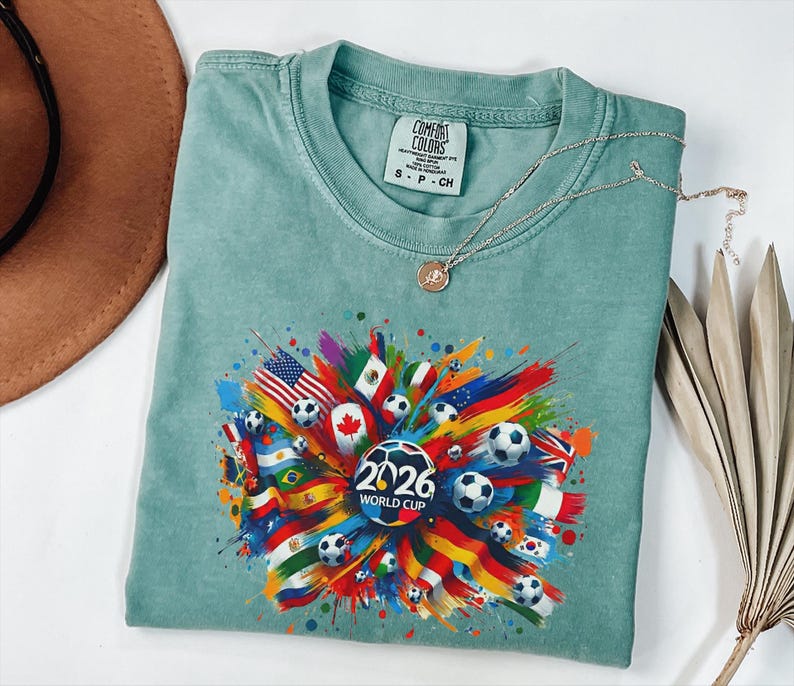 May include: A light teal Comfort Colors t-shirt featuring a colorful graphic design for the 2026 World Cup. The design includes soccer balls, flags from various countries, and the text "2026 WORLD CUP". A silver necklace with a pendant is also visible.