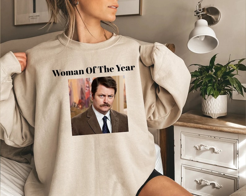 May include: A beige sweatshirt with the text "Woman Of The Year" and a picture of a man with a mustache and a suit.