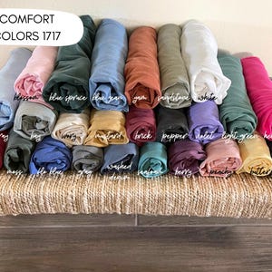 May include: A collection of rolled fabric in various colors, including blue, pink, and green, arranged on a woven surface. The text "COMFORT COLORS 1717" is at the top. Colors are labeled with names like "blossom" and "denim".