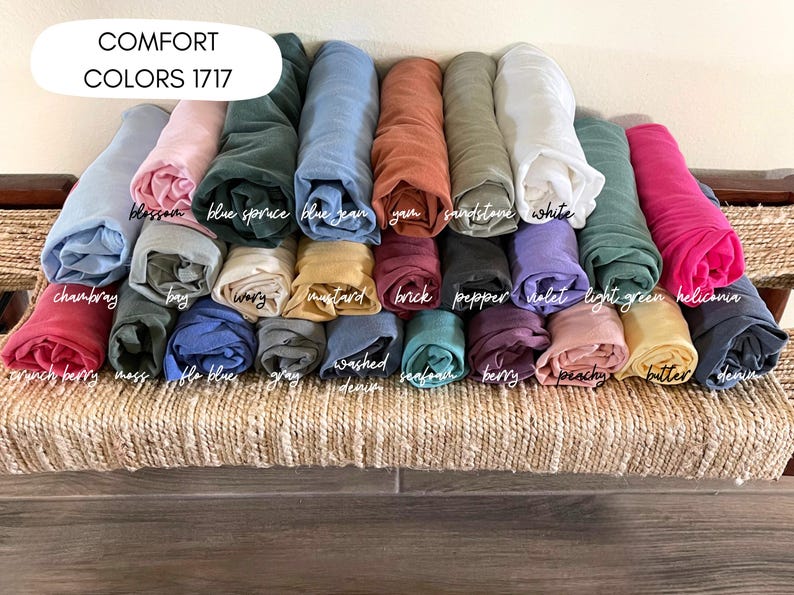 May include: A collection of rolled fabric in various colors, including blue, pink, and green, arranged on a woven surface. The text "COMFORT COLORS 1717" is at the top. Colors are labeled with names like "blossom" and "denim".
