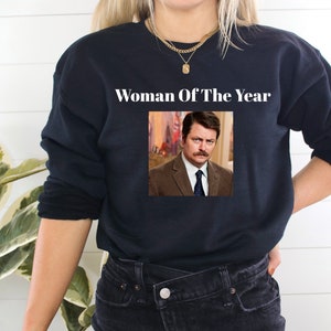 May include: Black sweatshirt with a white graphic that says "Woman Of The Year" and a picture of a man with a mustache.