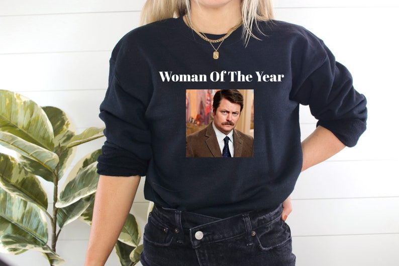 May include: Black sweatshirt with a white graphic that says "Woman Of The Year" and a picture of a man with a mustache.