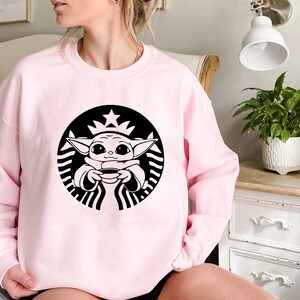 May include: Pink sweatshirt with a black and white graphic of a baby Yoda character holding a cup of coffee in a Starbucks logo.