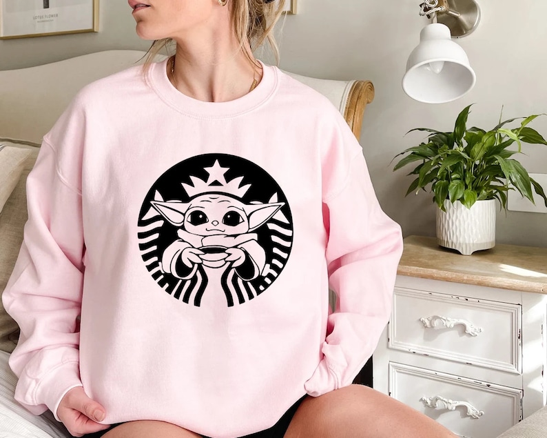 May include: Pink sweatshirt with a black and white graphic of a baby Yoda character holding a cup of coffee in a Starbucks logo.