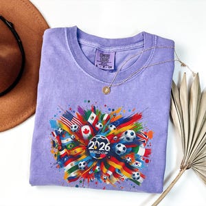 May include: A light purple t-shirt with a colorful graphic design featuring soccer balls, flags, and the text "2026 WORLD CUP". The design includes flags from various countries. The shirt is folded and displayed on a white surface.
