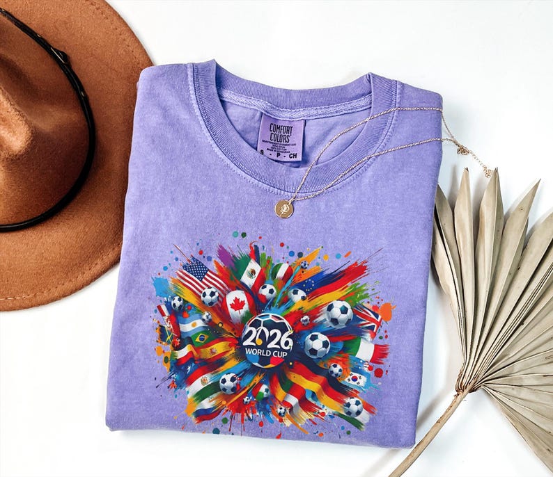 May include: A light purple t-shirt with a colorful graphic design featuring soccer balls, flags, and the text "2026 WORLD CUP". The design includes flags from various countries. The shirt is folded and displayed on a white surface.