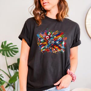 May include: Dark gray t-shirt featuring a colorful graphic design. The design includes soccer balls, flags from various countries, and the text "2026 WORLD CUP" in a circular emblem. The shirt has rolled-up sleeves.