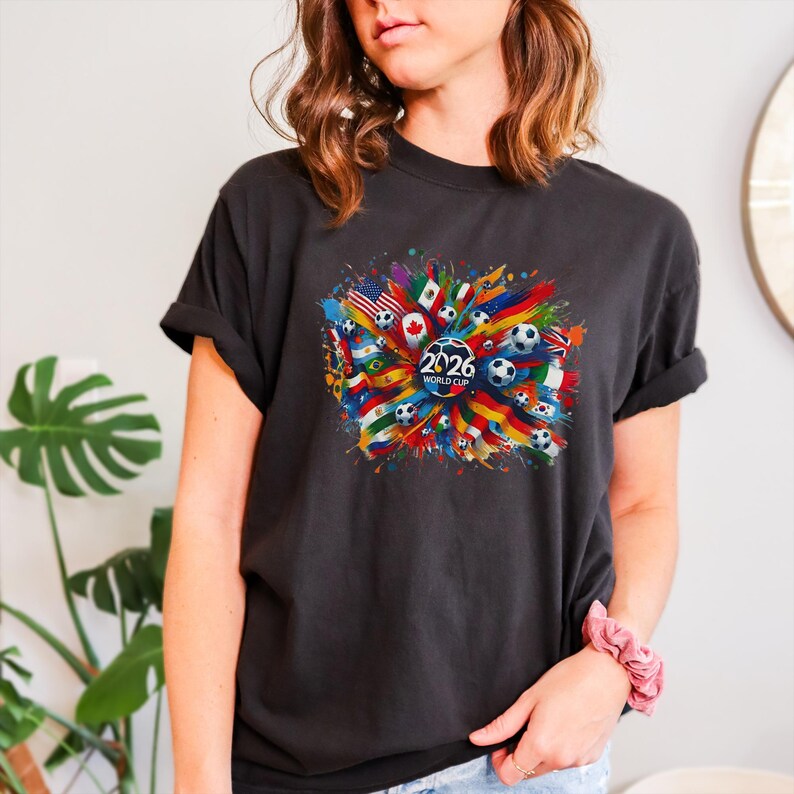 May include: Dark gray t-shirt featuring a colorful graphic design. The design includes soccer balls, flags from various countries, and the text "2026 WORLD CUP" in a circular emblem. The shirt has rolled-up sleeves.