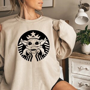 May include: A beige sweatshirt with a black and white graphic of a baby Yoda character holding a cup of coffee in a Starbucks logo.