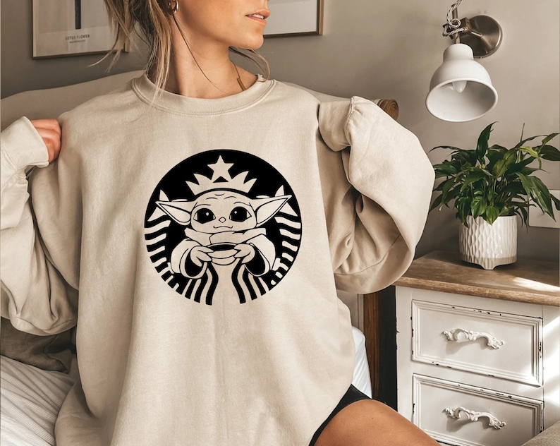 May include: A beige sweatshirt with a black and white graphic of a baby Yoda character holding a cup of coffee in a Starbucks logo.