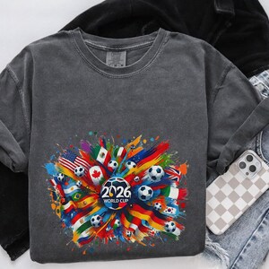 May include: A dark gray sweatshirt featuring a colorful graphic of soccer balls, flags, and the text "2026 WORLD CUP." The design includes flags from various countries. The sweatshirt is paired with jeans and a phone with a checkered case.