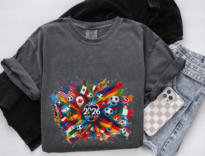 May include: A dark gray sweatshirt featuring a colorful graphic of soccer balls, flags, and the text "2026 WORLD CUP." The design includes flags from various countries. The sweatshirt is paired with jeans and a phone with a checkered case.