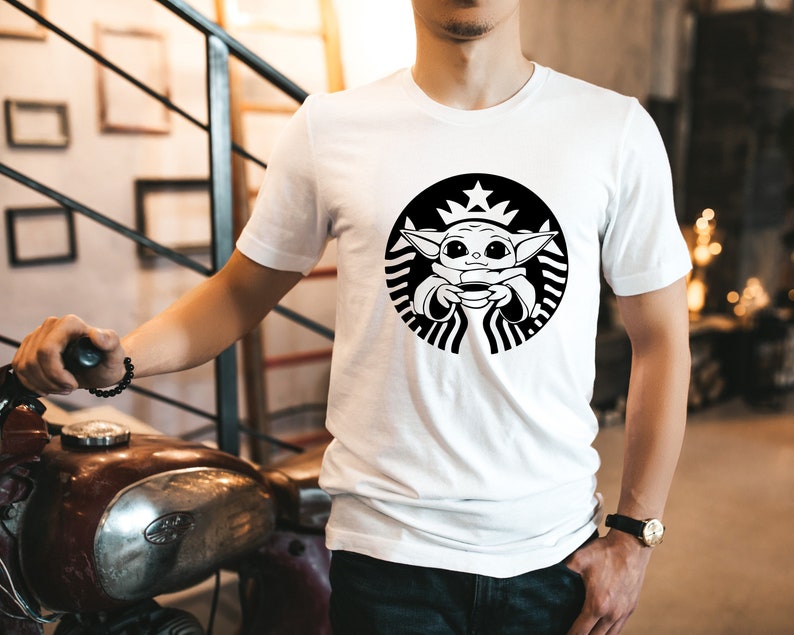 May include: White t-shirt with a black and white graphic of a cartoon character holding a cup. The graphic is a parody of the Starbucks logo.