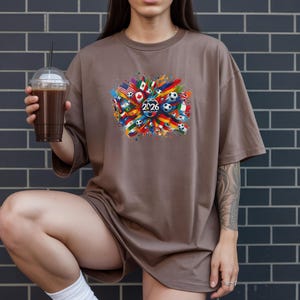 May include: Brown t-shirt with a colorful graphic featuring flags, soccer balls, and the text "2026". The design is centered on the shirt and includes a variety of national flags. The person is holding a clear plastic cup with a dark beverage.