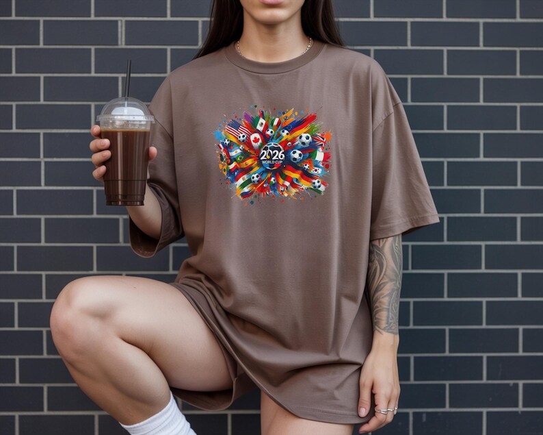 May include: Brown t-shirt with a colorful graphic featuring flags, soccer balls, and the text "2026". The design is centered on the shirt and includes a variety of national flags. The person is holding a clear plastic cup with a dark beverage.