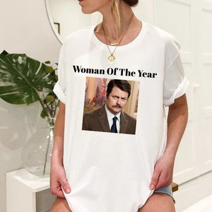 May include: White t-shirt with the text "Woman Of The Year" and a picture of a man with a mustache.