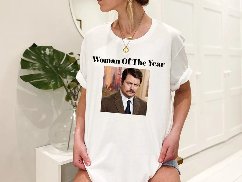 May include: White t-shirt with the text "Woman Of The Year" and a picture of a man with a mustache.