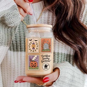 May include: A clear glass jar with a bamboo lid and a straw, featuring a Halloween-themed design. The design includes four postage stamp-style illustrations with a lantern, tombstone, and pumpkin. The text reads "Certified Spooky Gardener."