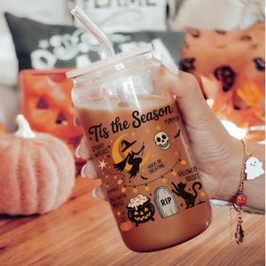 Peut inclure: Un gobelet en verre transparent avec un couvercle en bambou et une paille, rempli d'une boisson brune. Le gobelet est décoré de graphismes sur le thème d'Halloween et du texte "Tis the Season". Une citrouille et un bracelet à breloques sont en arrière-plan.