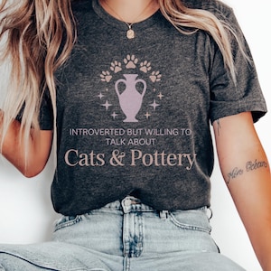 Puede incluir: Camiseta gris oscuro con el texto "INTROVERTED BUT WILLING TO TALK ABOUT Cats & Pottery". La camiseta presenta un gráfico rosa claro de un jarrón con huellas de patas y estrellas. La camiseta está hecha de un material suave.