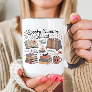 May include: White ceramic mug with a black handle and rim. The mug displays the text "Spooky Chapters Ahead" and Halloween illustrations such as books, a ghost, and a pumpkin.