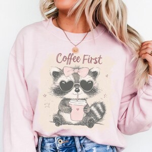 Könnte beinhalten: Rosafarbener Pullover mit einem Cartoon-Waschbären, der eine Sonnenbrille und eine Schleife trägt und aus einer Tasse trinkt. Der Text "Coffee First" ist auf dem Pullover aufgedruckt.