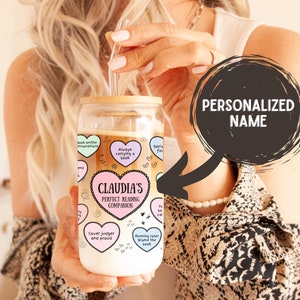 Peut inclure: Un gobelet en verre personnalisé avec un couvercle en bambou et une paille. Le gobelet a un design coloré avec des cœurs et du texte qui dit "Claudia's Perfect Reading Companion".
