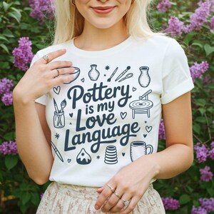 May include: White t-shirt with the text "Pottery is my Love Language" in navy blue, along with pottery-related illustrations. The shirt is worn with a beige lace skirt, set against a backdrop of purple flowers.