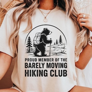 May include: Cream-colored t-shirt featuring a black graphic of a bear with a backpack and hiking stick, set against a mountain scene. The text reads "PROUD MEMBER OF THE BARELY MOVING HIKING CLUB."