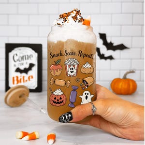 May include: A Halloween-themed glass of drink with whipped cream, sprinkles, and candy corn. The glass is decorated with Halloween treat illustrations and the words "Snack. Scare. Repeat." A sign in the background says "Come in for a Bite".