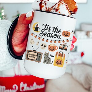 May include: White ceramic mug with a black handle, filled with hot chocolate and marshmallows. The mug is decorated with Halloween-themed illustrations, including pumpkins, ghosts, and the text "'tis the Season" and "Halloween Movies".