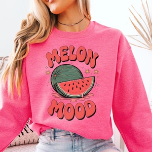 Puede incluir: Una sudadera rosa brillante con las palabras "MELON MOOD" en una fuente retro. El gráfico presenta una sandía verde y una rodaja de sandía. La sudadera es de estilo casual y cómodo.