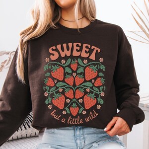 May include: A dark brown sweatshirt with a strawberry design. The design features red strawberries, green leaves, and the text "SWEET but a little wild" in a retro font. The sweatshirt is a crew neck style.