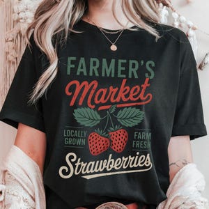 Puede incluir: Camiseta negra con un gráfico blanco que dice "Farmer's Market" en una fuente de estilo vintage. El gráfico también incluye dos fresas rojas con hojas verdes y el texto "Locally Grown" y "Farm Fresh" a cada lado de las fresas. El texto "Strawberries" está debajo de las fresas.