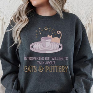 May include: Dark gray sweatshirt with the text "INTROVERTED BUT WILLING TO TALK ABOUT CATS & POTTERY". The graphic features a cat, a pottery wheel, and a small pot. The sweatshirt is a comfortable, casual style.