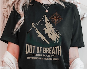 Op de afbeelding: Zwart T-shirt met een bergontwerp en de tekst "OUT OF BREATH - HIKING SOCIETY - DON'T WORRY, I'LL BE THERE IN A MINUTE." Het ontwerp bevat een kompas en een wandelaar. Het T-shirt heeft een ronde hals.