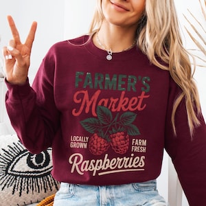 May include: Burgundy-colored sweatshirt with the text "Farmer's Market" in green and red, along with the words "Locally Grown" and "Farm Fresh." Below the text are two raspberry illustrations and the word "Raspberries."