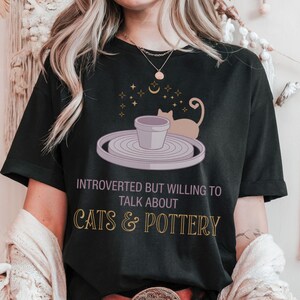 May include: Black t-shirt with a graphic featuring a cat, pottery wheel, and the text "Introverted but willing to talk about cats & pottery." The design includes stars and a crescent moon.