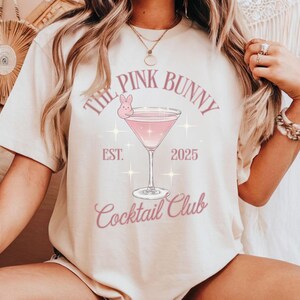 A trendy cocktail-themed t-shirt featuring a pink martini glass with a bunny accent and vintage lettering, perfect for bar lovers and aesthetic outfits. The main colors are pink and white.