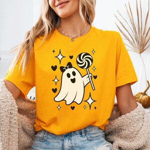 Puede incluir: Camiseta amarilla mostaza con un fantasma de dibujos animados sosteniendo un caramelo. El fantasma tiene un lazo negro, y el diseño incluye corazones y estrellas. La camiseta es de cuello redondo y manga corta.