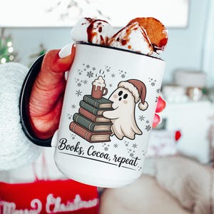 May include: White ceramic mug with a black rim and handle, filled with hot chocolate and topped with marshmallows and a biscuit. The mug features a ghost wearing a Santa hat, stacked books, snowflakes, and the text "Books, Cocoa, repeat."