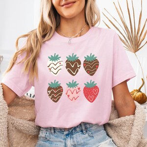 Puede incluir: Una camiseta rosa con un gráfico de seis fresas cubiertas de chocolate con corazones. Las fresas están dispuestas en dos filas de tres.