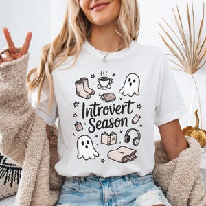 Puede incluir: Camiseta blanca con el texto "Introvert Season" y varias ilustraciones, incluyendo fantasmas, calcetines, una taza de café, un libro y auriculares. La camiseta se combina con pantalones cortos vaqueros.