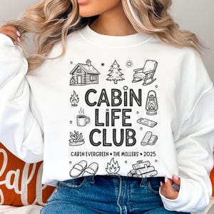 Könnte beinhalten: Weißes Sweatshirt mit schwarzem Aufdruck „CABIN LIFE CLUB“ und Illustrationen zum Thema Hütte. Der Text „CABIN EVERGREEN • THE MILLERS • 2025“ ist darunter gedruckt. Das Sweatshirt wird mit einer blauen Jeans getragen.
