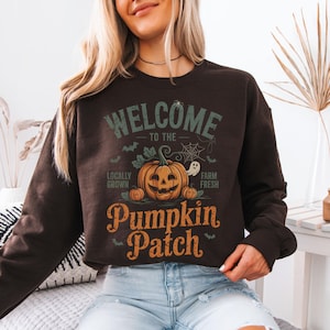 Peut inclure: Un sweat-shirt marron foncé avec l'inscription "WELCOME TO THE PUMPKIN PATCH" en orange et vert. Le motif comprend une citrouille d'Halloween, des chauves-souris, une toile d'araignée et un fantôme. Les mots "Locally Grown" et "Farm Fresh" sont également présents.